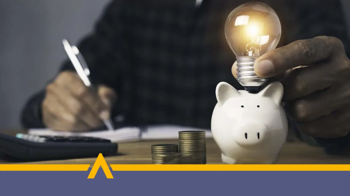 kobieta siedzącą przy stole z kalkulatorem i dokumentami, obok znajdują się skarbonka w kształcie świnki i żarówka